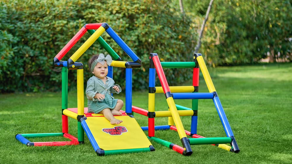 Baby seated in a low QUADRO playhouse beside a small climbing frame outdoors