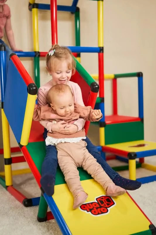 Two toddlers sliding together down a short QUADRO ramp indoors